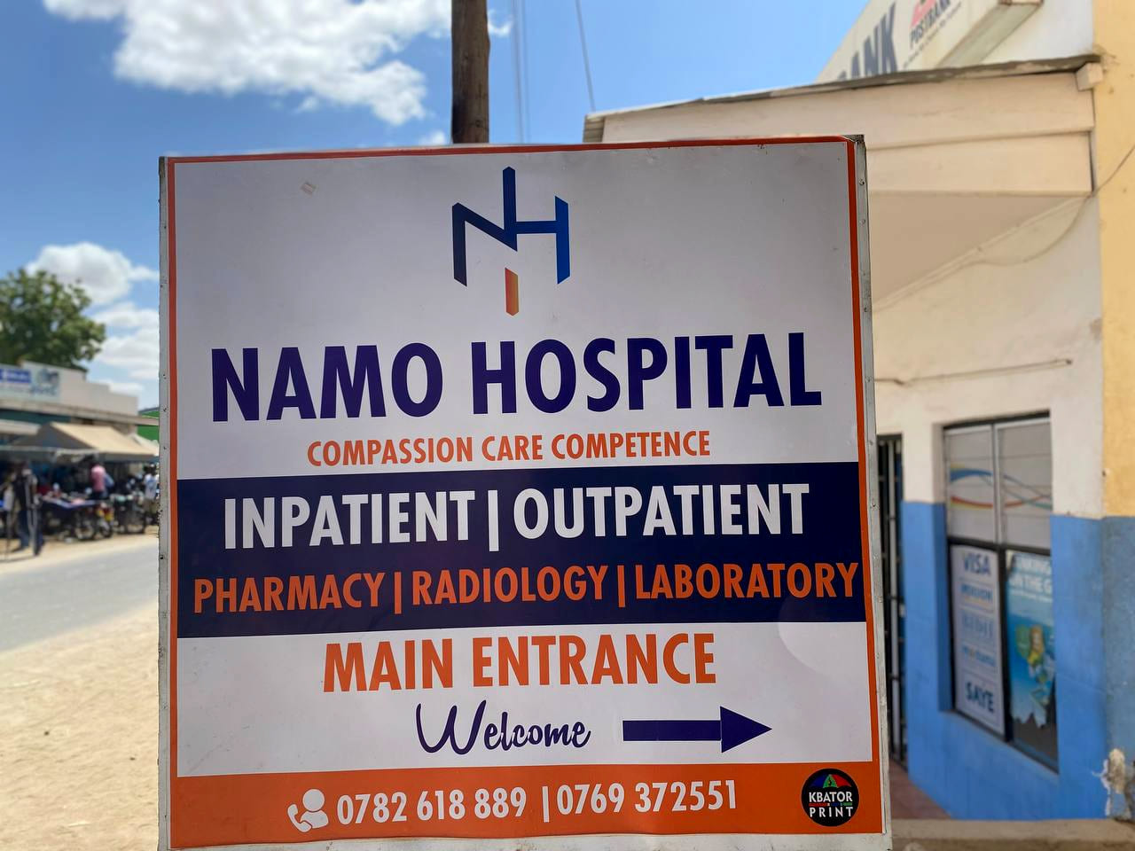 Namo Hospital's signboard on Equity Street in Lodwar, listing inpatient, outpatient, pharmacy, radiology and laboratory services, with an arrow to the main entrance. The PostBank building stands behind it.