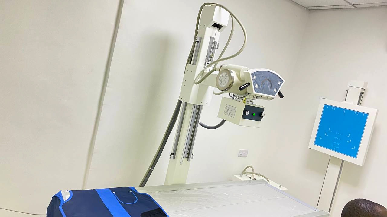 The digital X-ray room at Namo Hospital, with the X-ray tube mounted over the table, a lead apron folded on the table and a viewing screen alongside.