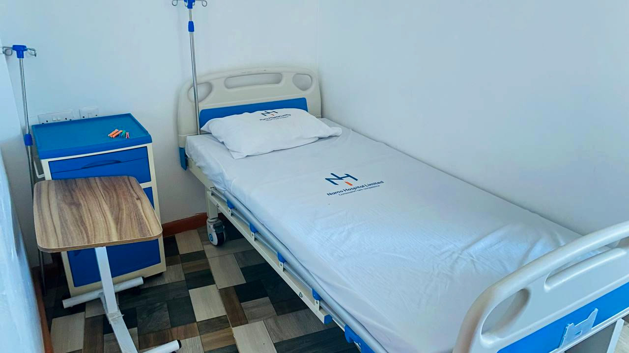 An inpatient bed at Namo Hospital made up with linen printed with the Namo Hospital Limited mark, alongside an overbed table and a bedside cabinet.