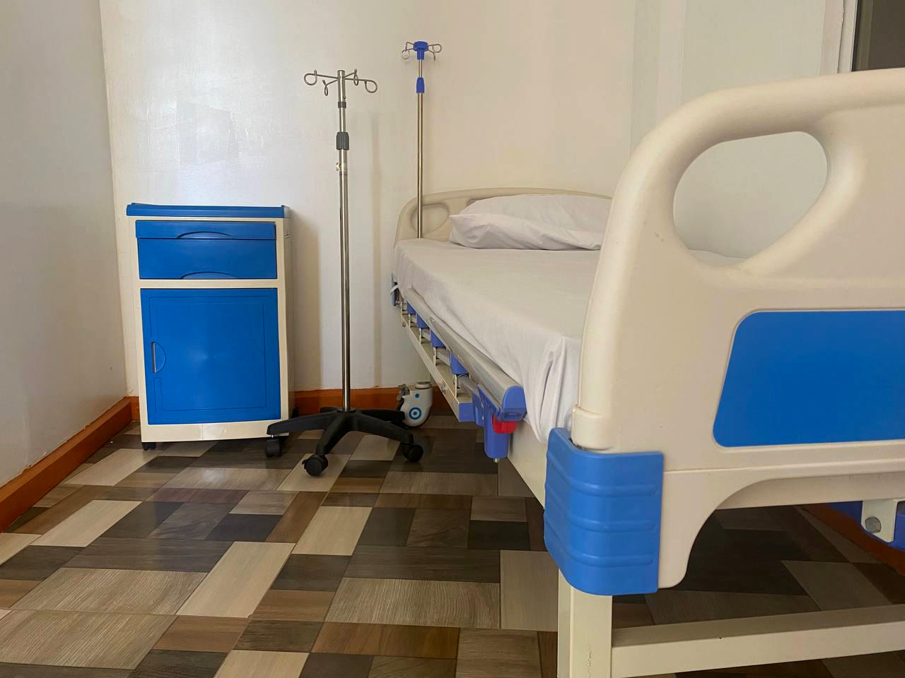 An inpatient bed at Namo Hospital with two intravenous stands and a bedside cabinet.