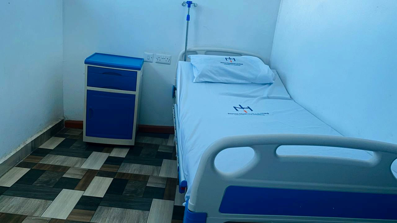 A hospital bed at Namo Hospital made up with branded sheets and pillowcase, with a bedside cabinet and an intravenous stand.