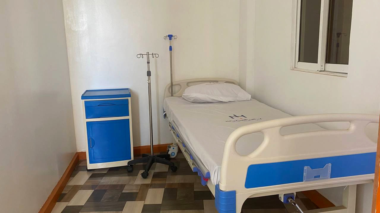 An inpatient room at Namo Hospital with a made bed beside a window, an intravenous stand and a bedside cabinet.